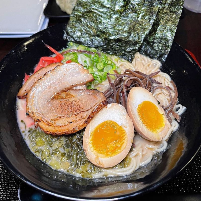A black bowl of Taro ramen with pork, soft-boiled eggs, green onions, and seaweed.