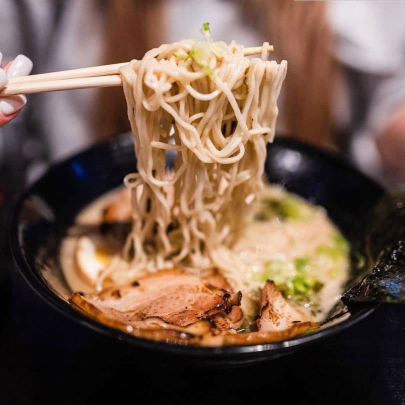 Chopsticks lifting a tangle of ramen noodles from a bowl with pork, egg, and scallions.
